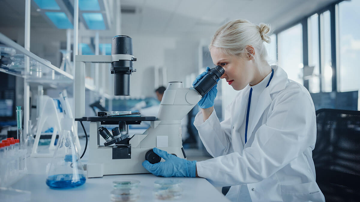 Portrait of female scientist looking under a microscope, analysing biochemical samples.